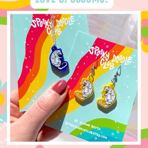 Pode incluir: Um colar e brincos com desenhos de gamb&aacute;. O colar tem um pingente de gamb&aacute; azul, e os brincos t&ecirc;m pingentes de gamb&aacute; amarelos. O texto "LOVE OPOSSUMS?" e "CHECK OUT MY OTHER OPOSSUM DESIGNS!" s&atilde;o vis&iacute;veis.