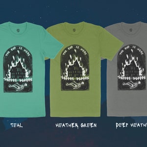 Op de afbeelding: Drie T-shirts met een grafische print van een brandend huis met een skelet dat ervoor ligt. De tekst "THE END IS HERE" is boven het huis gedrukt. De T-shirts zijn teal, heidegroen en diep heidekleurig.