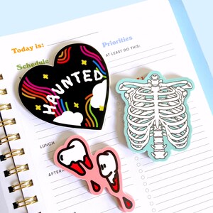 May include: Three colorful stickers with spooky themes. A black heart-shaped sticker with a rainbow design and the word "HAUNTED" in white letters. A blue sticker with a white skeleton ribcage. A pink sticker with two white teeth with red dripping blood.