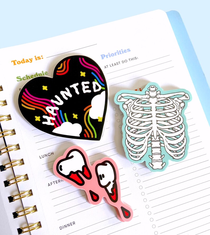 May include: Three colorful stickers with spooky themes. A black heart-shaped sticker with a rainbow design and the word "HAUNTED" in white letters. A blue sticker with a white skeleton ribcage. A pink sticker with two white teeth with red dripping blood.