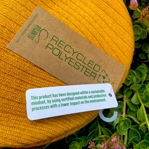 May include: A yellow knitted beanie with a brown tag that reads "RECYCLED POLYESTER" and a smaller white tag that says "This product has been designed within a sustainable mindset, by using certified materials and production processes with a lower impact on the environment."
