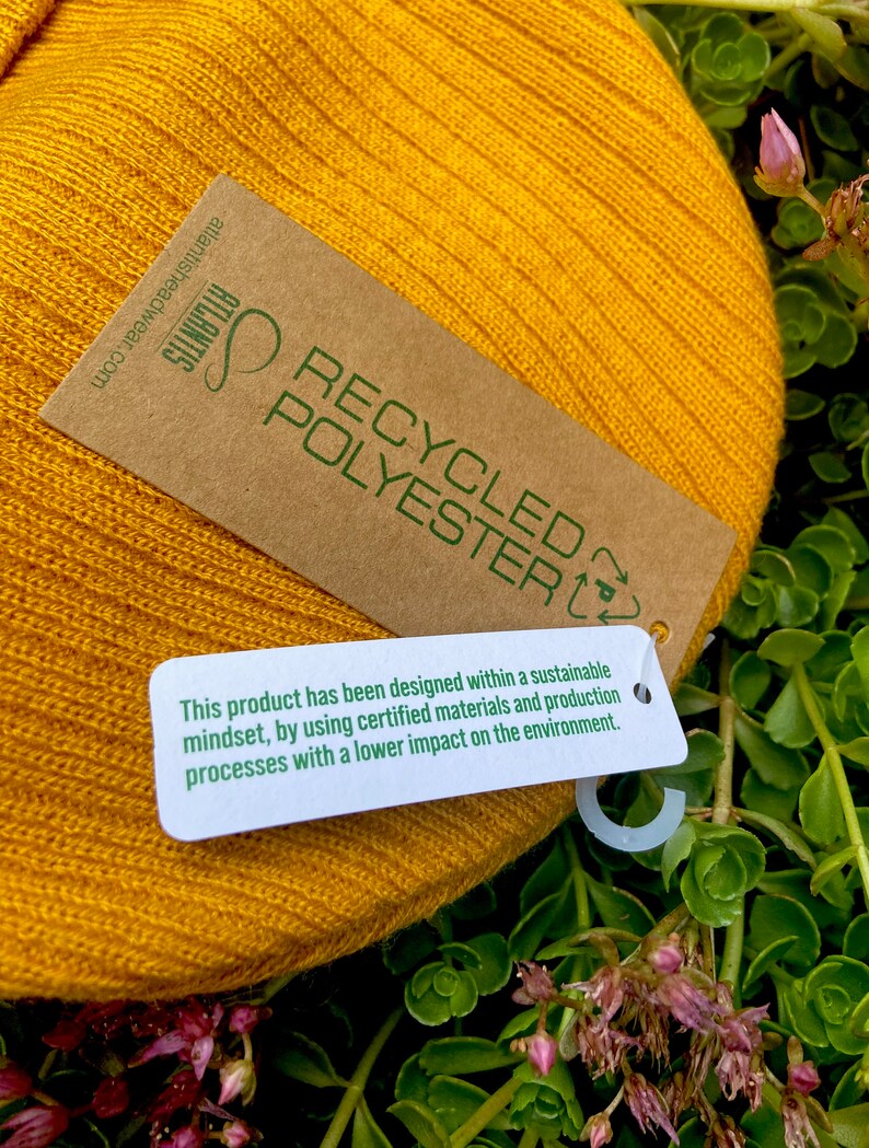 May include: A yellow knitted beanie with a brown tag that reads "RECYCLED POLYESTER" and a smaller white tag that says "This product has been designed within a sustainable mindset, by using certified materials and production processes with a lower impact on the environment."