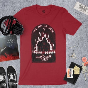 Op de afbeelding: Rood T-shirt met een witte afbeelding van een brandend huis met een skelet dat ervoor ligt. De tekst "The End Is Here" staat in een boog boven het huis.