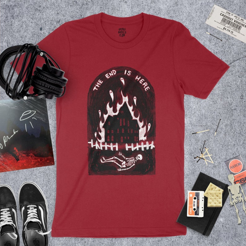 Op de afbeelding: Rood T-shirt met een witte afbeelding van een brandend huis met een skelet dat ervoor ligt. De tekst "The End Is Here" staat in een boog boven het huis.