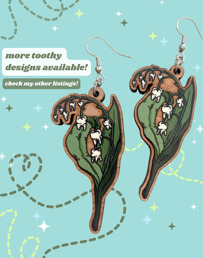 May include: A pair of wooden earrings featuring a whimsical design of lily of the valley flowers with teeth. The earrings are painted in shades of green and white, with a natural wood finish. The earrings are attached to silver hooks.
