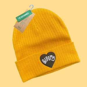 May include: A yellow beanie with a black heart-shaped patch that says "BLOOMY" in white lettering. The beanie is made from recycled polyester and is labeled "I'm Sustainable".