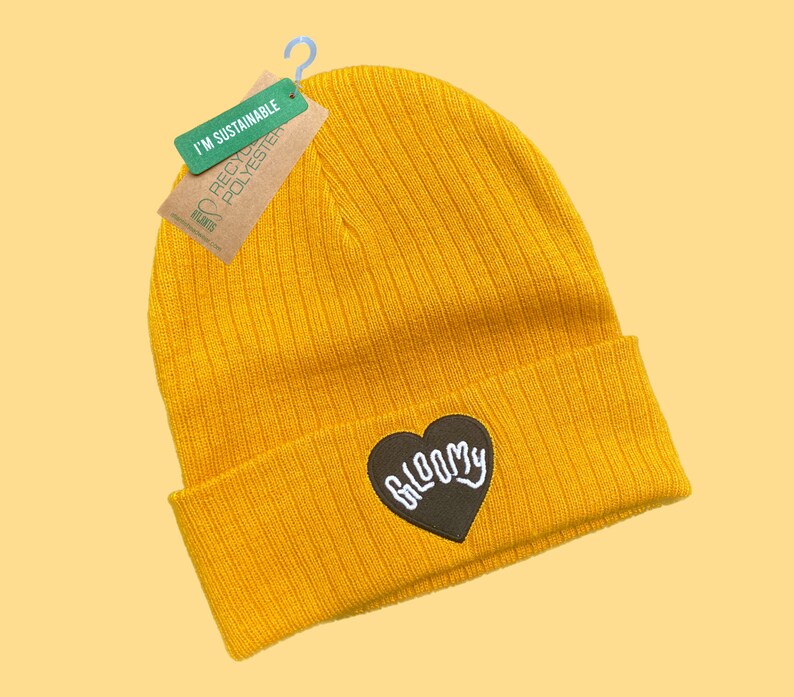 May include: A yellow beanie with a black heart-shaped patch that says "BLOOMY" in white lettering. The beanie is made from recycled polyester and is labeled "I'm Sustainable".