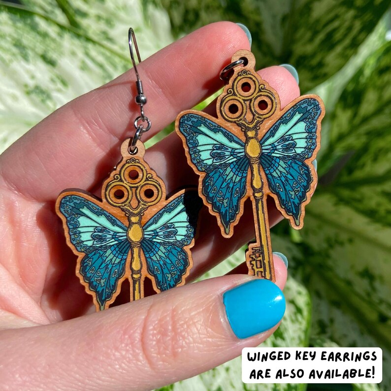 May include: A pair of wooden earrings shaped like keys with butterfly wings. The wings are blue and green with gold accents. The earrings are hanging from silver hooks.