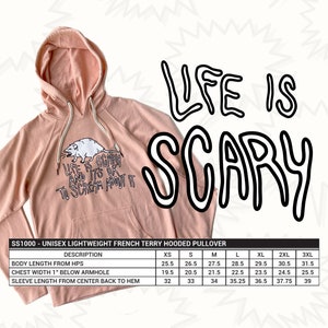 Pode incluir: Um moletom unissex rosa claro com capuz, bolso canguru e capuz com cord&atilde;o. A frente apresenta um gr&aacute;fico branco de um rato e o texto "LIFE IS SCARY AND IT'S OK TO SCREAM ABOUT IT". As palavras "LIFE IS SCARY" est&atilde;o escritas em letras pretas grandes.