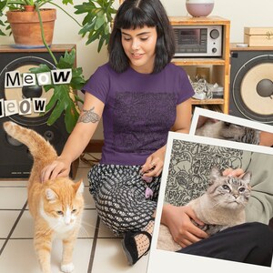 K&ouml;nnte beinhalten: Eine Frau tr&auml;gt ein lila T-Shirt mit einem schwarz-wei&szlig;en Katzendesign. Das Shirt tr&auml;gt die Aufschrift "Meow Meow" in schwarzen Buchstaben. Die Frau sitzt auf dem Boden mit einer Katze auf ihrem Scho&szlig;. Die Katze ist wei&szlig; und grau mit blauen Augen. Die Frau streichelt die Katze mit ihrer rechten Hand. Im Hintergrund ist eine zweite Katze zu sehen, eine orangefarbene Katze mit wei&szlig;en Flecken, die auf die Kamera zugeht.