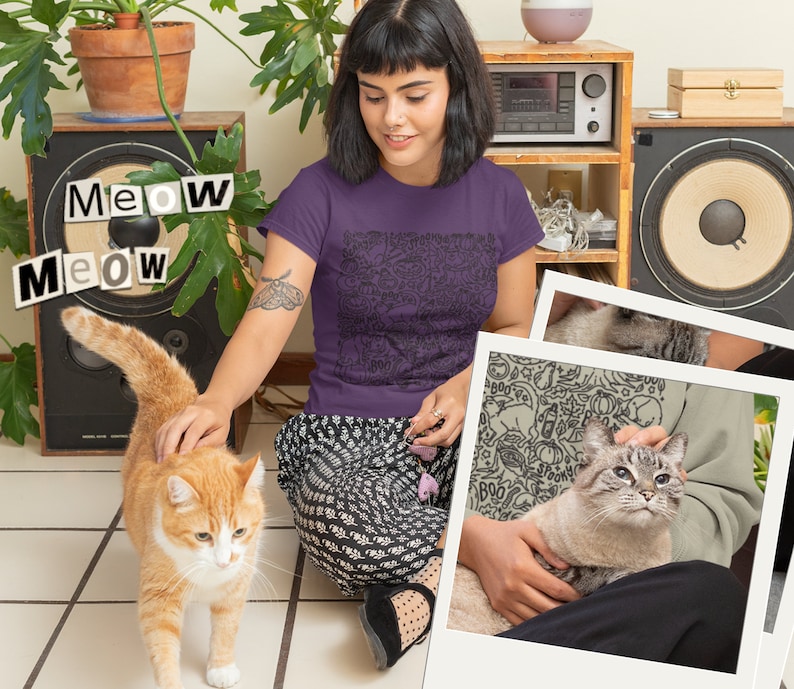 K&ouml;nnte beinhalten: Eine Frau tr&auml;gt ein lila T-Shirt mit einem schwarz-wei&szlig;en Katzendesign. Das Shirt tr&auml;gt die Aufschrift "Meow Meow" in schwarzen Buchstaben. Die Frau sitzt auf dem Boden mit einer Katze auf ihrem Scho&szlig;. Die Katze ist wei&szlig; und grau mit blauen Augen. Die Frau streichelt die Katze mit ihrer rechten Hand. Im Hintergrund ist eine zweite Katze zu sehen, eine orangefarbene Katze mit wei&szlig;en Flecken, die auf die Kamera zugeht.