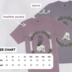 May include: Two t-shirts, one heather purple and one storm gray, featuring a gothic design. The design includes a raven perched on a skull, surrounded by candles, skeletal hands, and a leafy wreath. A size chart is also visible.