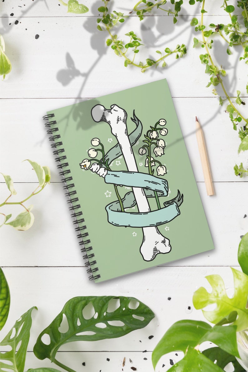 Op de afbeelding: Een groen spiraalnotitieblok met een illustratie van een wit bot en lelietjes-van-dalen bloemen. Het bot is omwikkeld met een blauw lint en er zijn witte sterren op de achtergrond.