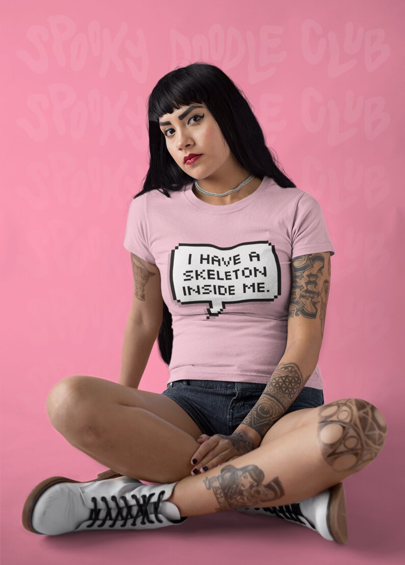 May include: A woman wearing a pink t-shirt with the text "I have a skeleton inside me." in a pixelated speech bubble. She is sitting with her legs crossed and wearing white boots.