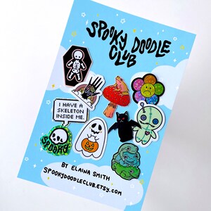 May include: A collection of 10 enamel pins with spooky and cute designs. The pins feature a skeleton, a hand with a googly eye, a frog on a mushroom, a flower with a sad face, a ghost, a bat, a voodoo doll, a skull, a pumpkin, and a stack of frogs. The pins are arranged on a blue background with white clouds and stars. The text "Spooky Doodle Club" is written in black at the top of the image. The text "By Elaina Smith Spookydoodleclub.etsy.com" is written in black at the bottom of the image.