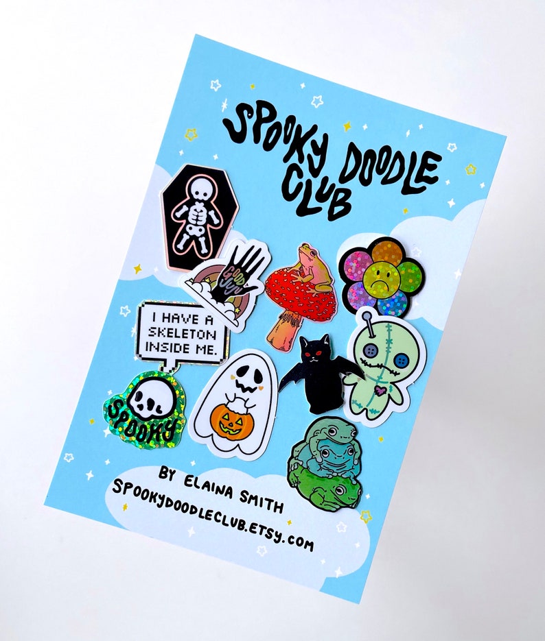 May include: A collection of 10 enamel pins with spooky and cute designs. The pins feature a skeleton, a hand with a googly eye, a frog on a mushroom, a flower with a sad face, a ghost, a bat, a voodoo doll, a skull, a pumpkin, and a stack of frogs. The pins are arranged on a blue background with white clouds and stars. The text "Spooky Doodle Club" is written in black at the top of the image. The text "By Elaina Smith Spookydoodleclub.etsy.com" is written in black at the bottom of the image.
