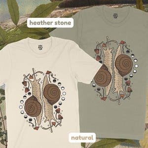 Pu&ograve; includere: Due magliette beige con una grafica di due lumache con funghi e fasi lunari. I testi "heather stone" e "natural" sono stampati sulle magliette.