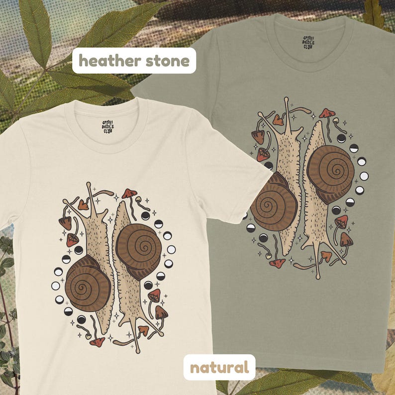 Pu&ograve; includere: Due magliette beige con una grafica di due lumache con funghi e fasi lunari. I testi "heather stone" e "natural" sono stampati sulle magliette.