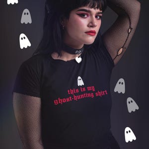 Op de afbeelding: Een zwart T-shirt met de tekst "this is my ghost-hunting shirt" in rode gotische letters. Het shirt wordt gedragen door een persoon met zwart haar en donkere make-up. Er zijn verschillende witte geestillustraties rond de persoon.