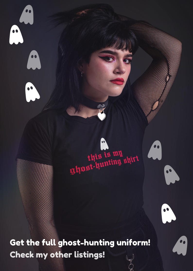 Op de afbeelding: Een zwart T-shirt met de tekst "this is my ghost-hunting shirt" in rode gotische letters. Het shirt wordt gedragen door een persoon met zwart haar en donkere make-up. Er zijn verschillende witte geestillustraties rond de persoon.