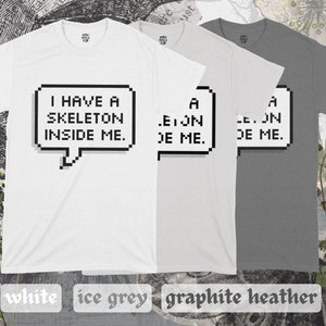 May include: Three t-shirts in white, light gray, and dark gray with a pixelated speech bubble graphic that says "I have a skeleton inside me."