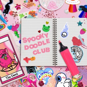 May include: A pink notebook with the words "Spooky Doodle Club" written on it. The notebook is surrounded by various stickers, including a pink fuzzy creature, a green frog, a skull, a rat, and a rainbow UFO.