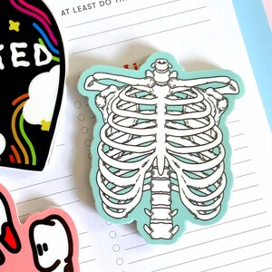 May include: A sticker of a white ribcage on a light blue background. The sticker is on a lined sheet of paper with the text "Priorities" and "At Least Do This:" at the top of the page.