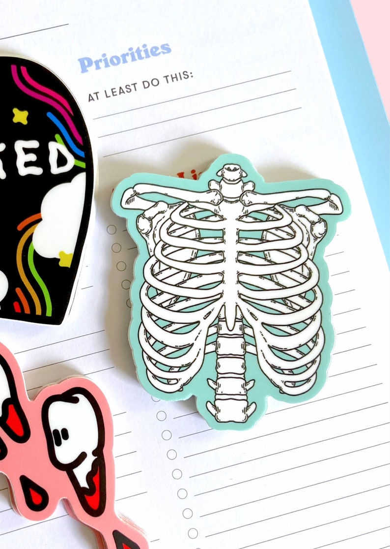 May include: A sticker of a white ribcage on a light blue background. The sticker is on a lined sheet of paper with the text "Priorities" and "At Least Do This:" at the top of the page.