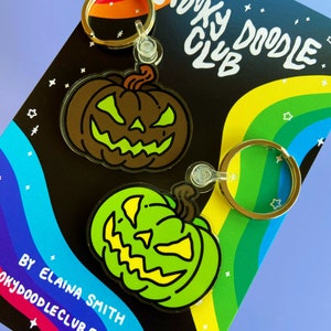 May include: Two green and brown Halloween pumpkin keychains with a spooky doodle club logo on a rainbow background. The keychains are made of acrylic and have a black outline. The text on the card reads "Spooky Doodle Club" and "By Elaina Smith Spookydoodleclub.etsy.com".