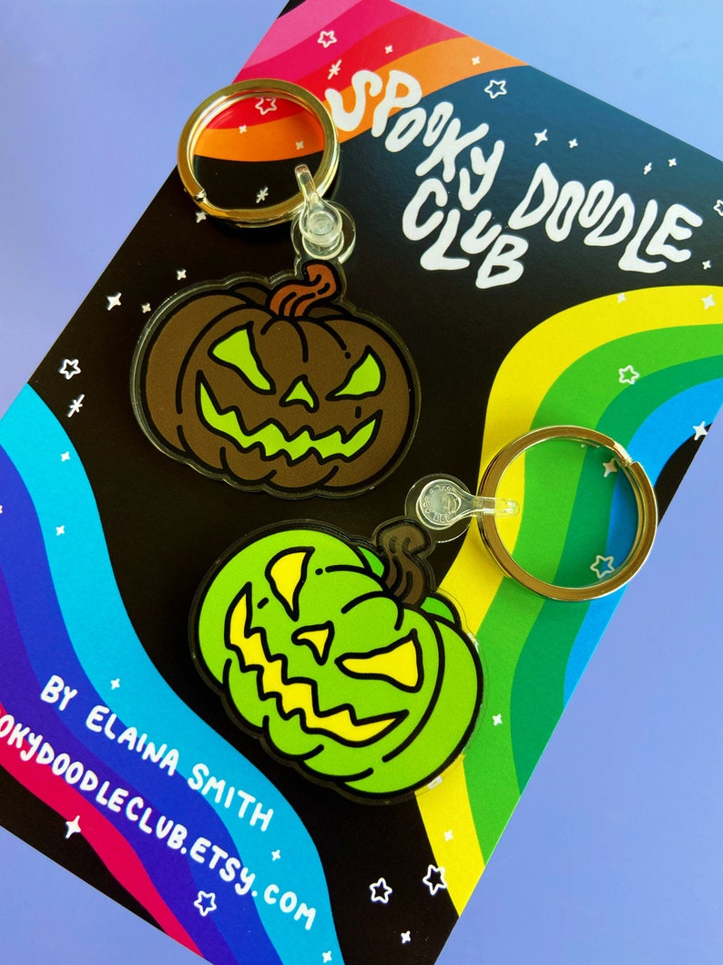 May include: Two green and brown Halloween pumpkin keychains with a spooky doodle club logo on a rainbow background. The keychains are made of acrylic and have a black outline. The text on the card reads "Spooky Doodle Club" and "By Elaina Smith Spookydoodleclub.etsy.com".