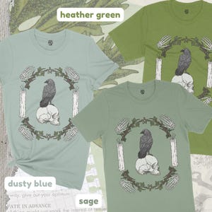 May include: Three short-sleeved t-shirts in dusty blue, heather green, and sage. Each shirt features a graphic of a raven perched on a skull, surrounded by a wreath, candles, and skeletal hands. The text "heather green", "dusty blue", and "sage" are visible.