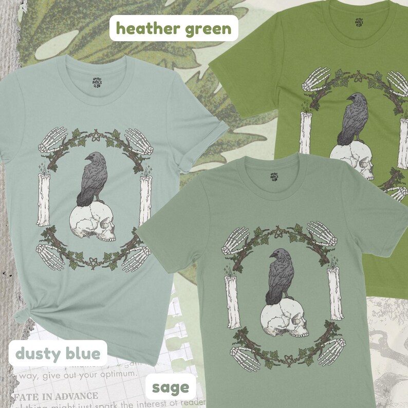 May include: Three short-sleeved t-shirts in dusty blue, heather green, and sage. Each shirt features a graphic of a raven perched on a skull, surrounded by a wreath, candles, and skeletal hands. The text "heather green", "dusty blue", and "sage" are visible.