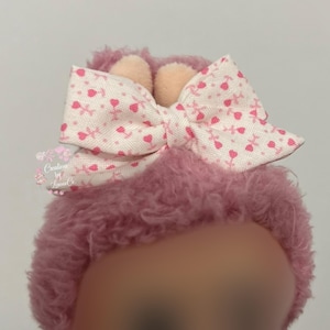 May include: A pink, fluffy headband with a large bow. The bow is white with a pattern of pink hearts and flowers. Two small, peach-colored ears are attached to the headband.