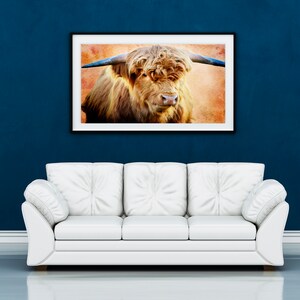 May include: A framed print of a Highland cow hangs above a white sofa against a dark blue wall. The artwork features a close-up of the cow's face, showcasing its long, shaggy brown fur and large horns. The background has a textured, orange hue.