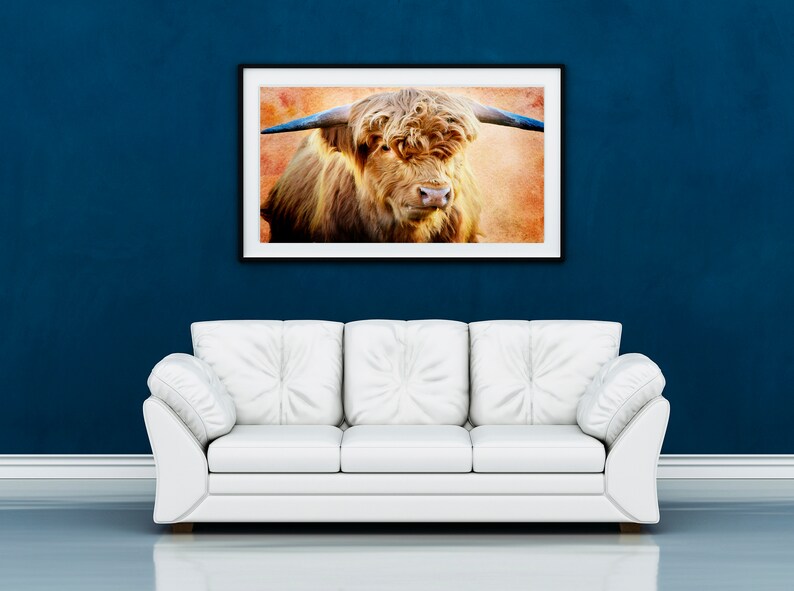 May include: A framed print of a Highland cow hangs above a white sofa against a dark blue wall. The artwork features a close-up of the cow's face, showcasing its long, shaggy brown fur and large horns. The background has a textured, orange hue.