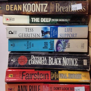 May include: A stack of hardcover books with various titles and authors, including James Patterson, Dean Koontz, and Patricia Cornwell. The books are in different colors, with visible text and labels.