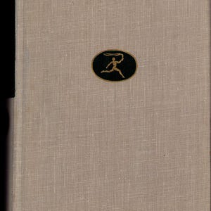 May include: A hardcover book with a beige linen cover. A black oval emblem with a gold figure holding a flowing object is centered on the cover. The book's spine is a darker shade of beige.