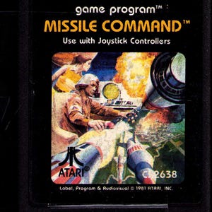 May include: A vintage Atari Missile Command game cartridge. The label features a colorful illustration of a military scene with explosions and a soldier. Text includes "MISSILE COMMAND" and "Use with Joystick Controllers". The Atari logo is visible.