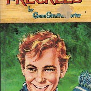 以下が含まれることがあります： ジーン・ストラットン＝ポーター著「FRECKLES」というタイトルのヴィンテージ本の表紙。赤い髪とそばかすのある笑顔の若い人が、青と白のストライプのスカーフを身につけています。背景は緑色です。