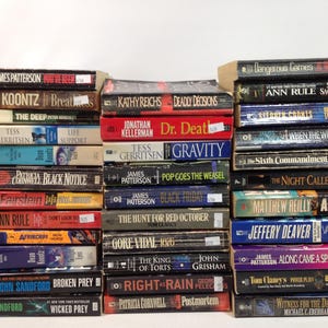 May include: A stack of paperback books with various titles and authors. The spines display titles like "Broken Prey" by John Sandford, "Along Came a Spider" by James Patterson, and "Deadly Decisions" by Kathy Reichs. The books are arranged in neat rows.