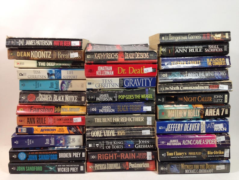 May include: A stack of paperback books with various titles and authors. The spines display titles like "Broken Prey" by John Sandford, "Along Came a Spider" by James Patterson, and "Deadly Decisions" by Kathy Reichs. The books are arranged in neat rows.
