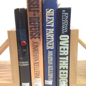 Könnte beinhalten: Vier gebundene Bücher von Jonathan Kellerman, darunter "Time Bomb", "Self-Defense", "Silent Partner" und "Over the Edge", werden von einem hölzernen Buchstützen gehalten. Die Bücher haben verschiedene Farben und sind vertikal angeordnet.