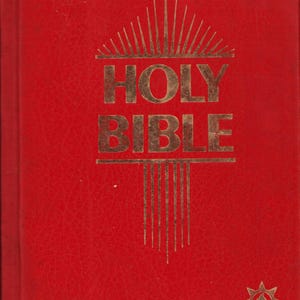 May include: A red hardback book with the words "HOLY BIBLE" in gold lettering. The cover has a textured appearance and a gold sunburst design above the title. A gold starburst design is in the lower right corner.
