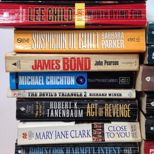 May include: A stack of paperback books with various titles and authors. The books have colorful covers, including titles like "Power Down," "Lee Child," and "Sphere." The spines display the authors' names and book titles.
