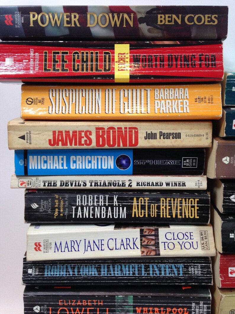 May include: A stack of paperback books with various titles and authors. The books have colorful covers, including titles like "Power Down," "Lee Child," and "Sphere." The spines display the authors' names and book titles.