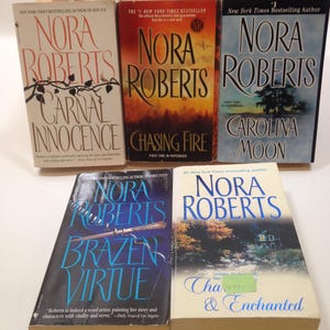 Op de afbeelding: Een verzameling van vijf paperback boeken van auteur Nora Roberts. De boeken zijn verticaal gestapeld, met de titels "Carnal Innocence", "Chasing Fire", "Carolina Moon", "Brazen Virtue" en "Charmed & Enchanted".