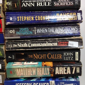 May include: A stack of paperback books with titles including "Dangerous Games", "Small Sacrifices", "Saucer", "When the Wind Blows", and "Area 7". The books have various colors and fonts, with some featuring author names and price tags.