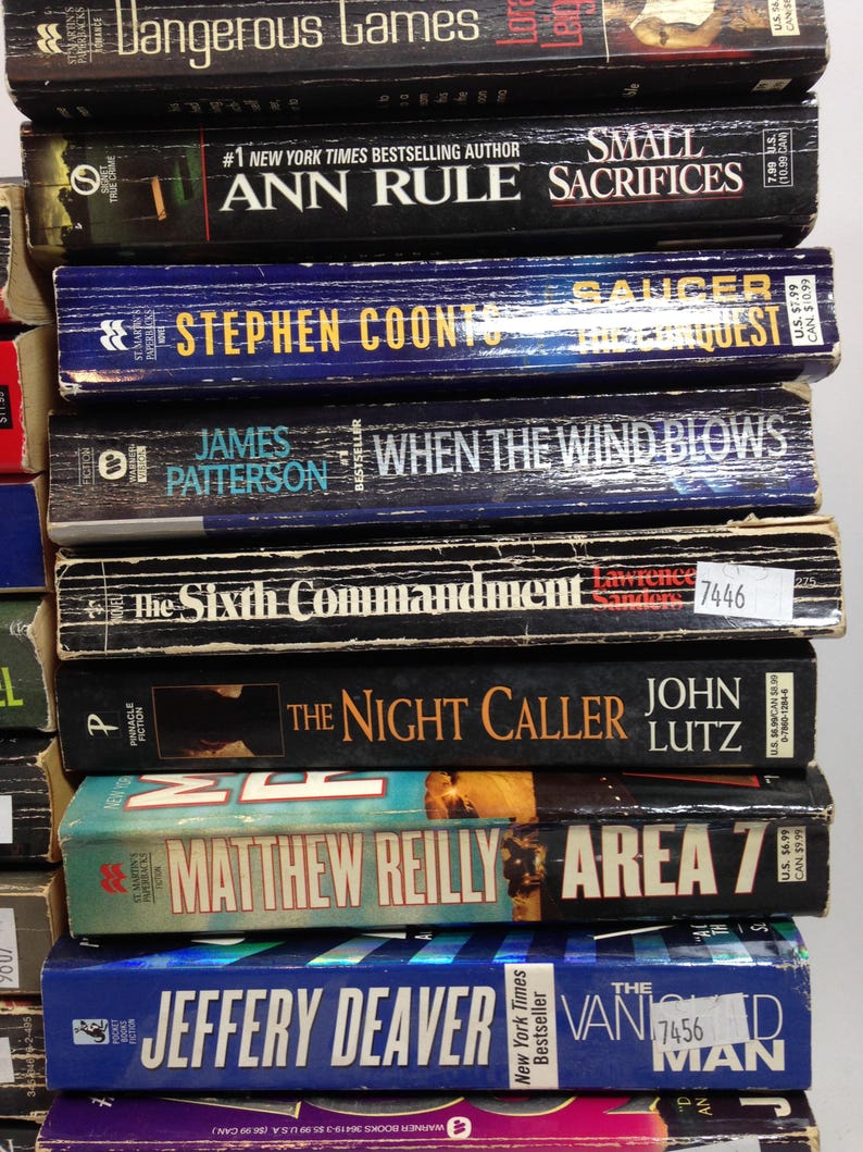 May include: A stack of paperback books with titles including "Dangerous Games", "Small Sacrifices", "Saucer", "When the Wind Blows", and "Area 7". The books have various colors and fonts, with some featuring author names and price tags.