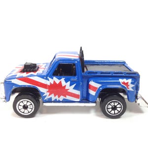 May include: A blue toy pickup truck with red and white starburst and stripe designs. The truck has black wheels and a black air intake on the hood. The truck bed is also blue.