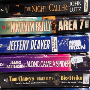 May include: A stack of paperback books with titles including "The Night Caller," "Area 7," and "Along Came a Spider." The spines display author names and book titles in various fonts and colors, with price stickers visible.